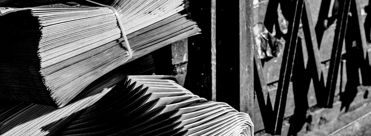 Two neatly bundles of neatly stacked newspapers on ground outside beside metal gate, in black and white