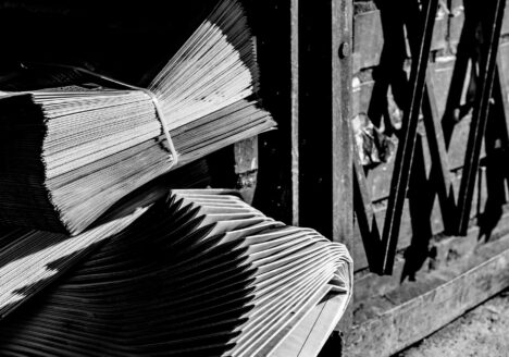 Two neatly bundles of neatly stacked newspapers on ground outside beside metal gate, in black and white