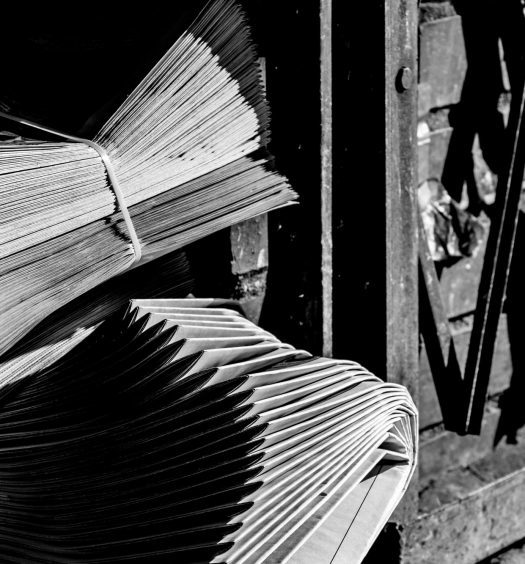 Two neatly bundles of neatly stacked newspapers on ground outside beside metal gate, in black and white