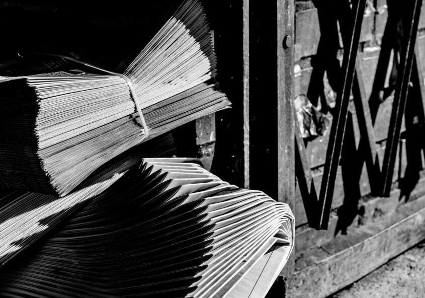 Two neatly bundles of neatly stacked newspapers on ground outside beside metal gate, in black and white