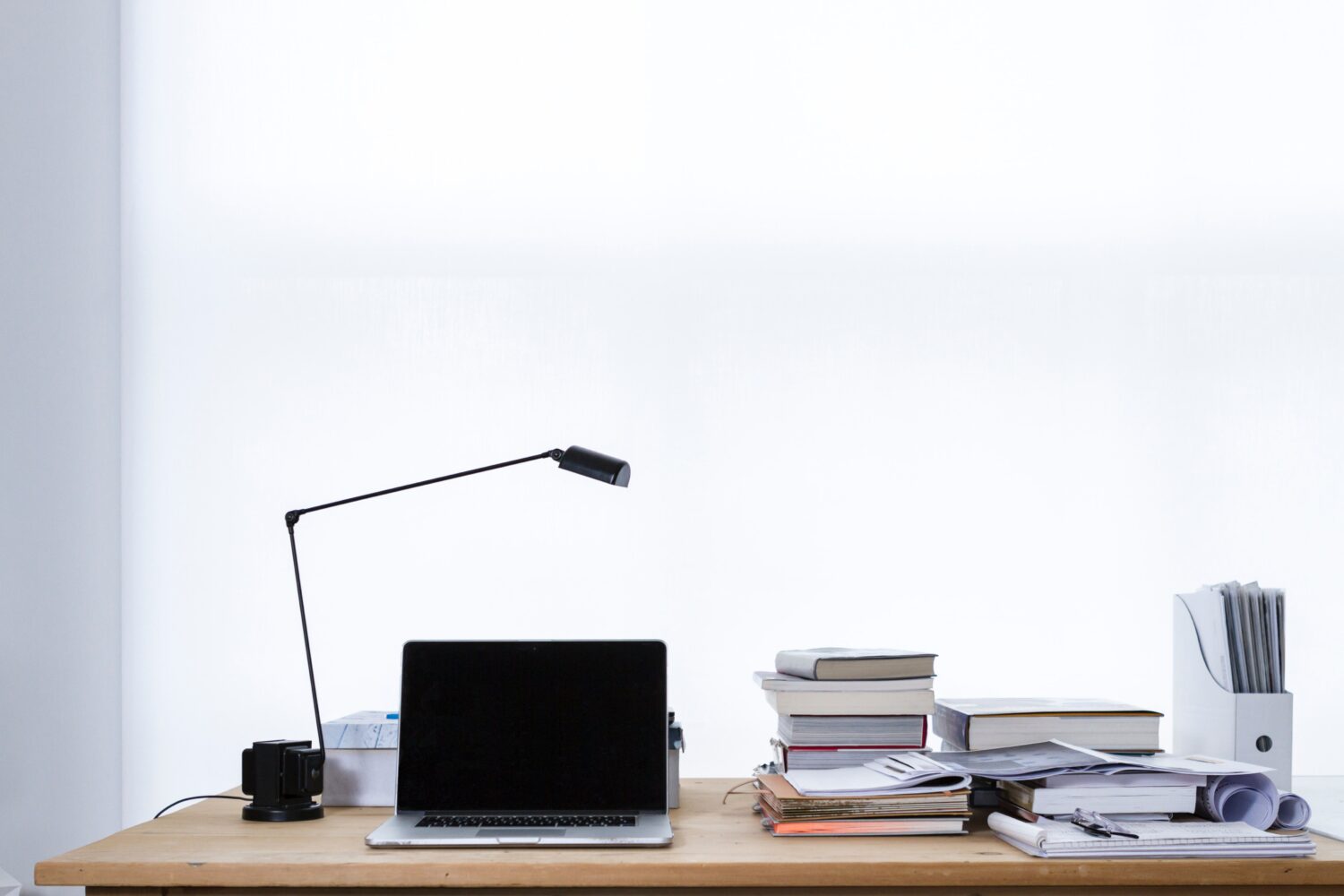 Laptop on wood desk with a lamp, stacks of books and folders in front of a white wall.