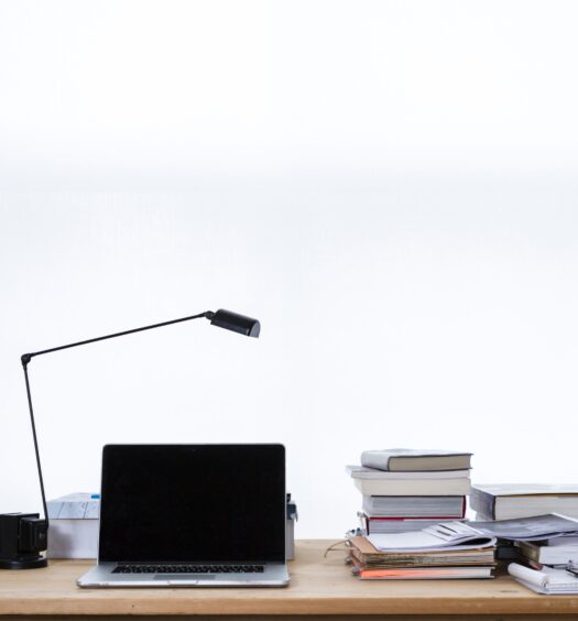 Laptop on wood desk with a lamp, stacks of books and folders in front of a white wall.