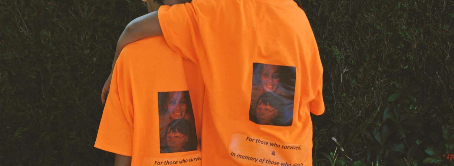 Two children, one with arm around the other facing away, wearing orange t-shirts that read "For those who survived & in memory of those who didn't"