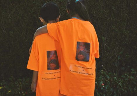Two children, one with arm around the other facing away, wearing orange t-shirts that read "For those who survived & in memory of those who didn't"