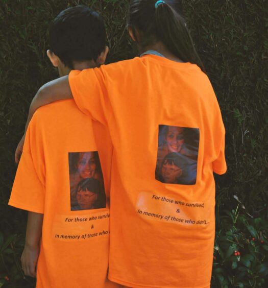 Two children, one with arm around the other facing away, wearing orange t-shirts that read "For those who survived & in memory of those who didn't"