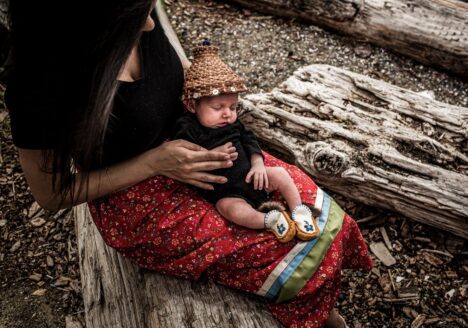 A woman sits outside on a tree log holding an infant.