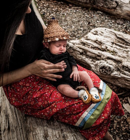 A woman sits outside on a tree log holding an infant.