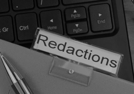 File labeled "redacted" sitting on top of a computer keyboard. The tip of a pen is laying on on file in bottom left corner.