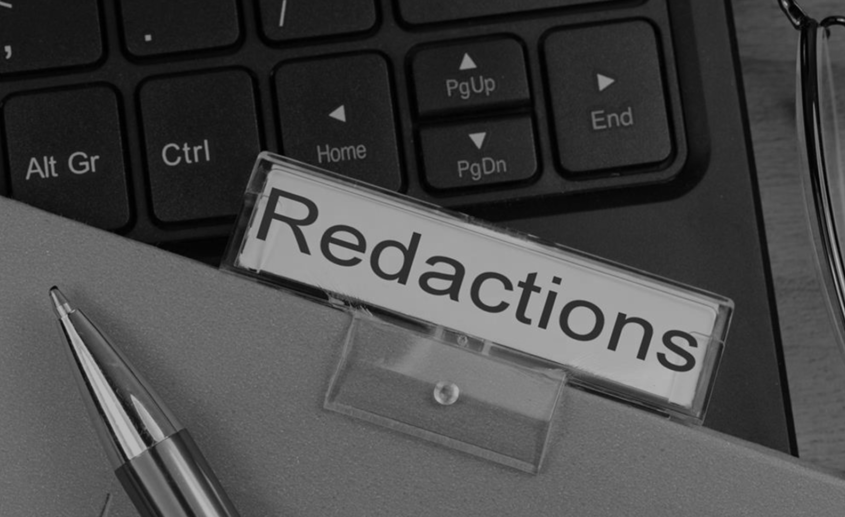 File labeled "redacted" sitting on top of a computer keyboard. The tip of a pen is laying on on file in bottom left corner.