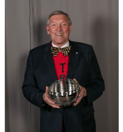 John Honderich in a red t-shirt, bowtie and blazer smiling and holding his Canadian Journalism Foundation Lifetime Achievement Award
