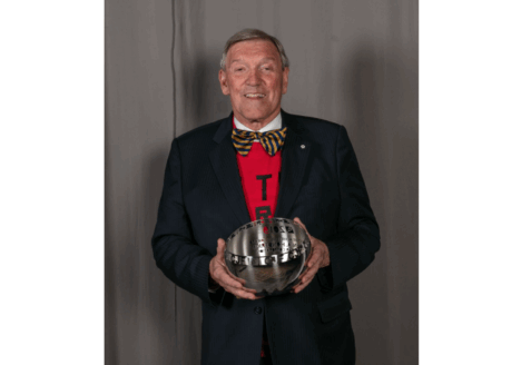 John Honderich in a red t-shirt, bowtie and blazer smiling and holding his Canadian Journalism Foundation Lifetime Achievement Award