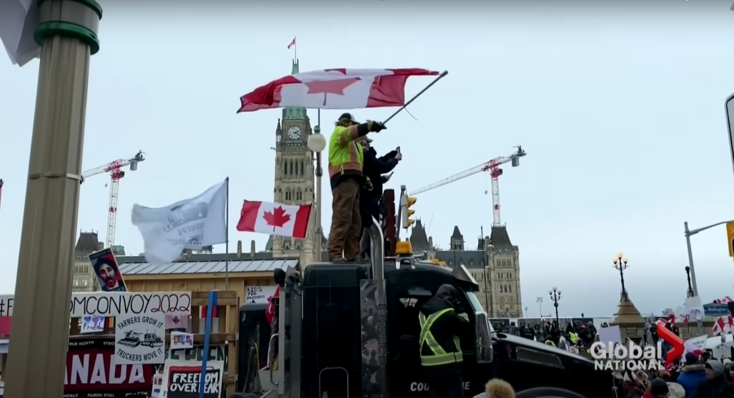 Protesters waving Canadian flag stand on top of and surrounding trucks in Ottawa