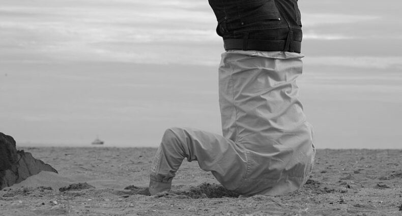 A person upside down with their head buried in sand on a beach. Photo in black and white