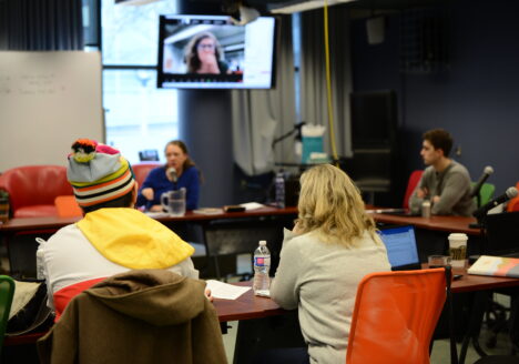 Four people sit around a set of tables facing each other. One person appears on a monitor overhead.