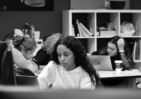 Four students work at laptops, one in foreground and three in background. Photo in black and white