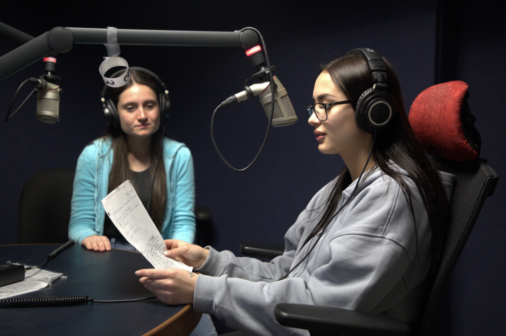 Two students in a podcast studio facing mics