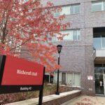 Red Richcraft Hall sign with black posts, outside of building. Tree with red fall leaves behind the sign.