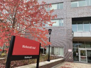 Red Richcraft Hall sign with black posts, outside of building. Tree with red fall leaves behind the sign.