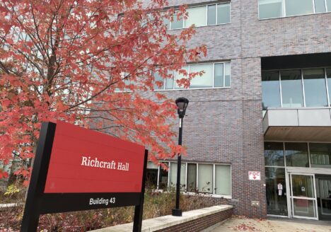 Red Richcraft Hall sign with black posts, outside of building. Tree with red fall leaves behind the sign.