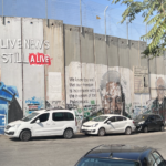 This is a picture of the Bethlehem side of the wall that separates the West Bank from Israel. On the wall is a painting of slain journalist, Shireen Abu Akleh, a Palestinian-American journalist who was killed by Israeli soldiers while wearing a blue press jacket and reporting on a raid at a refugee camp in Jenin. In the picture Shireen is seen wearing her blue, press jacket. He brown-blonde hair is straight and shoulder-length. Behind her is a graffiti text that says, Live News Still Alive.