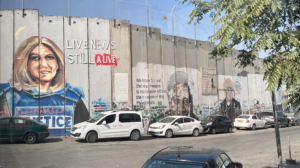 This is a picture of the Bethlehem side of the wall that separates the West Bank from Israel. On the wall is a painting of slain journalist, Shireen Abu Akleh, a Palestinian-American journalist who was killed by Israeli soldiers while wearing a blue press jacket and reporting on a raid at a refugee camp in Jenin. In the picture Shireen is seen wearing her blue, press jacket. He brown-blonde hair is straight and shoulder-length. Behind her is a graffiti text that says, Live News Still Alive.