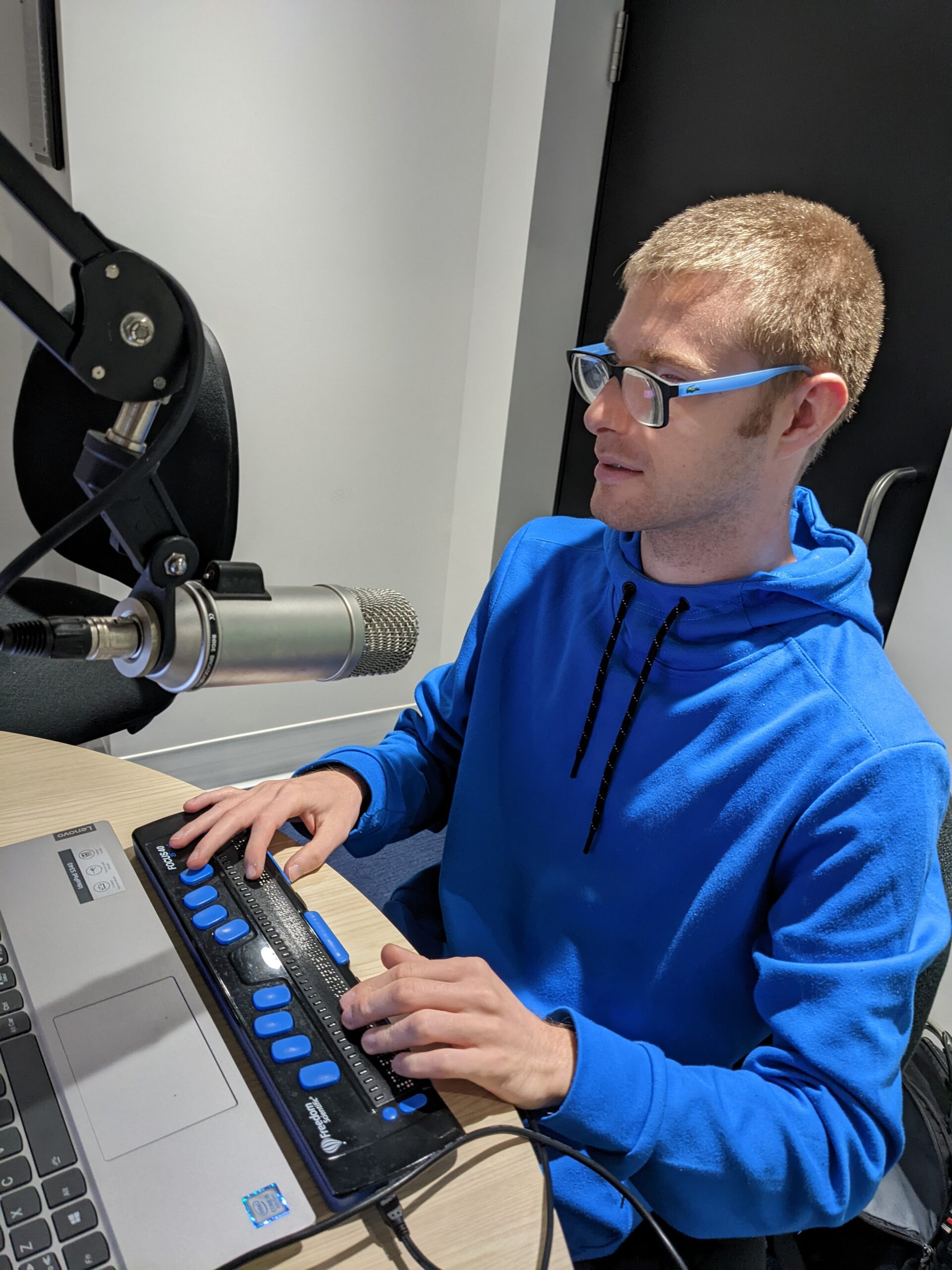 In this image, Benjamin is sitting and working. He is wearing a blue sweatshirt and glasses with a blue frame. 