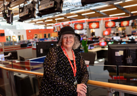 Cathy Browne is standing against a bannister in front of the CBC newsroom. She is wearing a printed black blouse and a black hat, and a red lanyard around her neck.