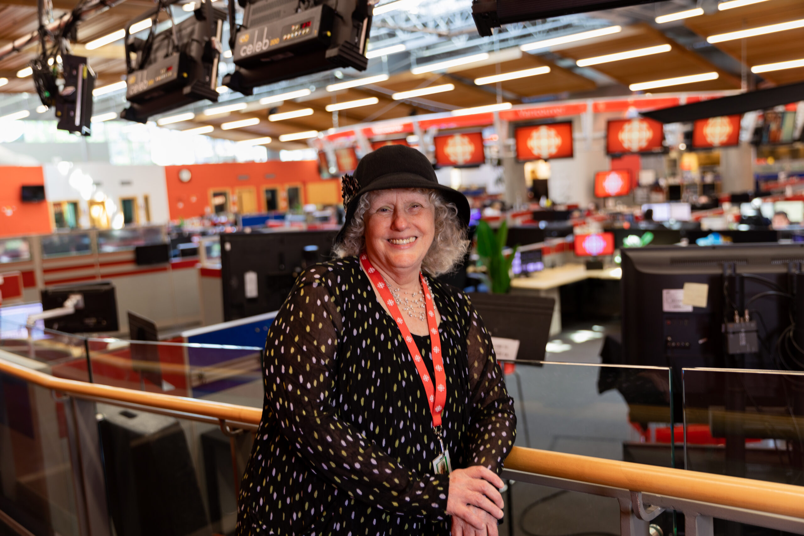 Cathy Browne is standing against a bannister in front of the CBC newsroom. She is wearing a printed black blouse and a black hat, and a red lanyard around her neck.