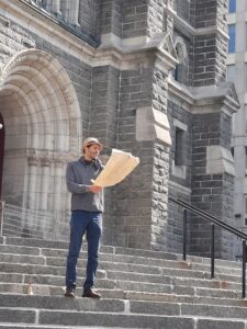 Charles Auguste is standing on the steps of an old building, wearing a straw hat and reading from a large piece of paper