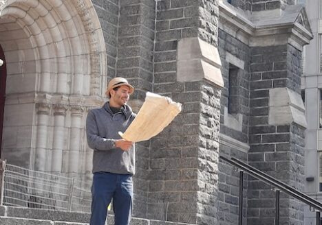 Charles Auguste is standing on the steps of an old building, wearing a straw hat and reading from a large piece of paper