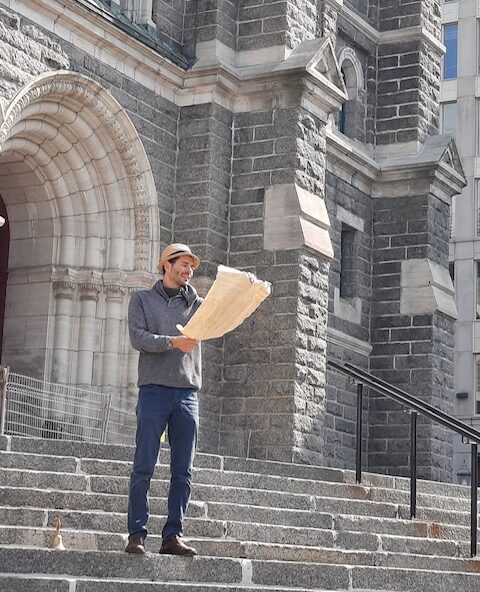 Charles Auguste is standing on the steps of an old building, wearing a straw hat and reading from a large piece of paper