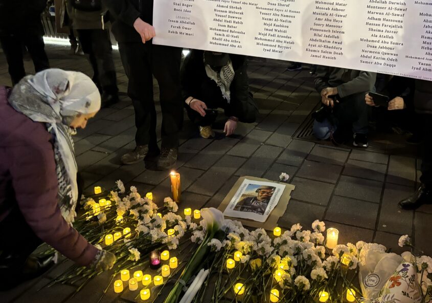 In this photo a woman is putting a flower down at a vigil for slain Palestininan, Lebanese and Israeli journalists. She is bending down in front of a poster carried by two people with all the journalists' names on it.