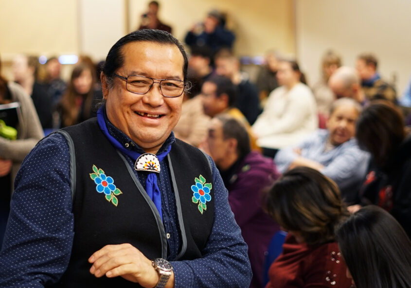 In this picture William Greenland is captured in a room full of people. He is wearing a black sleeveless vest with blue flowers on either side. He is wearing a blue long-sleeved shirt and is wearing a watch on his left wrist. Greeland has his black hair tied back and is wearing black-rimmed glases.