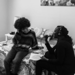 In this picture Garvey Bailey is sitting on the right side of the image holding a mic and interviewing singer Madisan McFerrin who is dressed in a sweater and jeans and sitting on a bed next to her with one hand in her hair and the other on the bed behind her.
