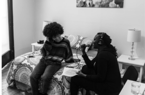 In this picture Garvey Bailey is sitting on the right side of the image holding a mic and interviewing singer Madisan McFerrin who is dressed in a sweater and jeans and sitting on a bed next to her with one hand in her hair and the other on the bed behind her.