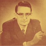 In this picture Tom Kent is wearing a suit and tie and sitting facing the camera. He is wearing glasses and his hair is brushed with a side margin.