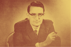 In this picture Tom Kent is wearing a suit and tie and sitting facing the camera. He is wearing glasses and his hair is brushed with a side margin.