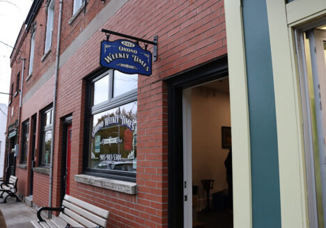 Exterior of the Orono Weekly Times. Brick facade with window and blue sign.
