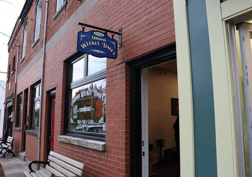 Exterior of the Orono Weekly Times. Brick facade with window and blue sign.