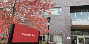 Red Richcraft Hall sign with black posts, outside of building. Tree with red fall leaves behind the sign.